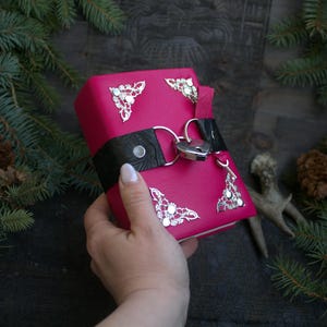 May include: A hot pink leather-bound journal with silver filigree corner accents and a heart-shaped lock closure. The journal has a black leather strap and is held in a hand.