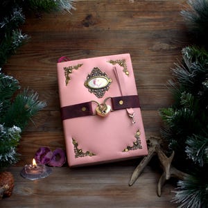 May include: Pink leather journal with a decorative lock and key closure. The journal features ornate corner accents and a dark brown strap. A decorative oval emblem is centered on the cover. The journal is set on a wooden surface with pine branches.