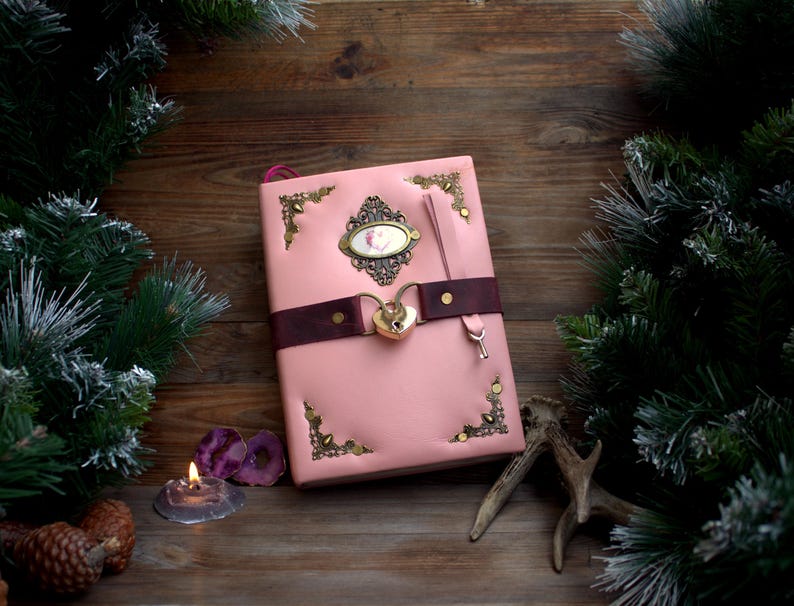 May include: Pink leather journal with a decorative lock and key closure. The journal features ornate corner accents and a dark brown strap. A decorative oval emblem is centered on the cover. The journal is set on a wooden surface with pine branches.