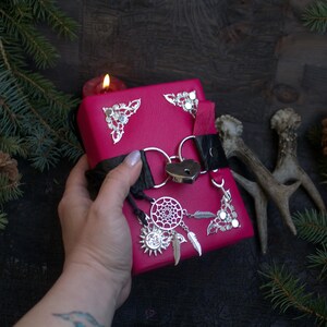 May include: A magenta-colored journal with silver metal corner accents and a heart-shaped lock. A black leather strap secures the journal, which features a dreamcatcher charm and feather pendants. The journal is held by a hand.