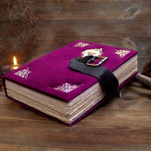 May include: A deep purple journal with a black leather strap and buckle. The cover is adorned with silver decorative elements and a framed picture. The aged pages suggest a vintage or antique style.