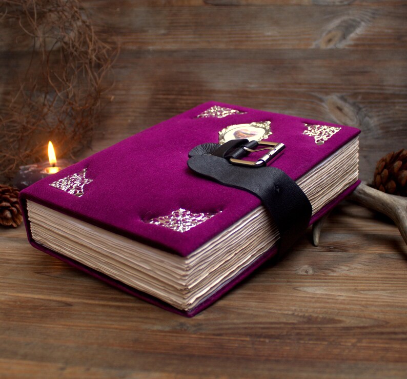 May include: A deep purple journal with a black leather strap and buckle. The cover is adorned with silver decorative elements and a framed picture. The aged pages suggest a vintage or antique style.