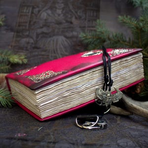 May include: A handmade, red leather-bound journal with ornate gold detailing on the cover. A black cord with a moth pendant hangs from the journal. The journal is surrounded by pine branches and a candle.