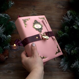 May include: A pink journal with a decorative brass lock and corner accents. The journal has a dark burgundy ribbon closure and a small oval embellishment with a floral design. The journal is held in a person's hand.