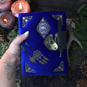 May include: A royal blue velvet journal featuring ornate brass corner protectors, a central oval cabochon displaying a triple moon symbol, and a black leather strap closure secured with a brass padlock and key. Two black ribbons hang with a star charm.