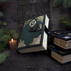 May include: A dark green, leather-bound book with gold filigree corners and a decorative clasp. The book has a black strap closure and a small framed image on the cover. A lit candle and evergreen branches are in the background.