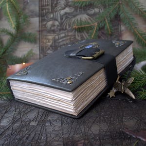 May include: A dark gray leather-bound journal featuring a black strap closure with a brass-toned buckle. The cover is adorned with ornate silver-toned metal corner accents and a central emblem. The pages are thick, off-white, and deckled-edged.
