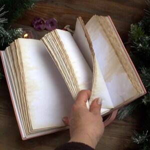 May include: An open, antique-style journal with aged, cream-colored pages. The journal has a pink cover and a pink ribbon bookmark. The pages are slightly yellowed, giving it a vintage look. The journal is surrounded by Christmas decorations.
