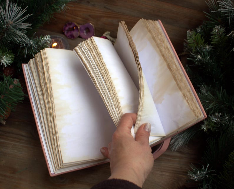 May include: An open, antique-style journal with aged, cream-colored pages. The journal has a pink cover and a pink ribbon bookmark. The pages are slightly yellowed, giving it a vintage look. The journal is surrounded by Christmas decorations.