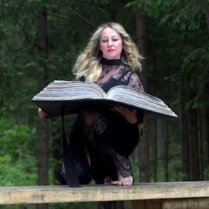 May include: A large, thick, open book with numerous textured, off-white pages held by a person wearing a black lace garment with a high collar. The book has a dark, solid cover and a long black ribbon bookmark hanging down from the bottom edge.