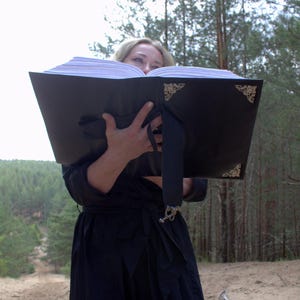 May include: A large, black book with ornate corner designs is held open, revealing white pages. A black ribbon with a decorative clasp is attached. The book is held in front of a person in a black dress, with a forest background.
