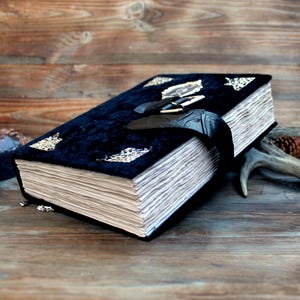 May include: A large, dark blue velvet book with gold leaf accents and a black leather strap closure. The book's pages are aged and textured, suggesting a vintage or antique style. The book is on a wooden surface.