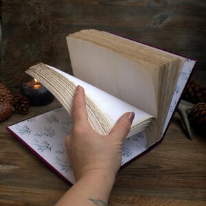 May include: An open book with aged, cream-coloured pages. The book has a deep purple cover and a floral patterned interior. A hand is turning the pages, revealing the texture of the paper. The background includes a wooden surface, pine cones, and a lit candle.