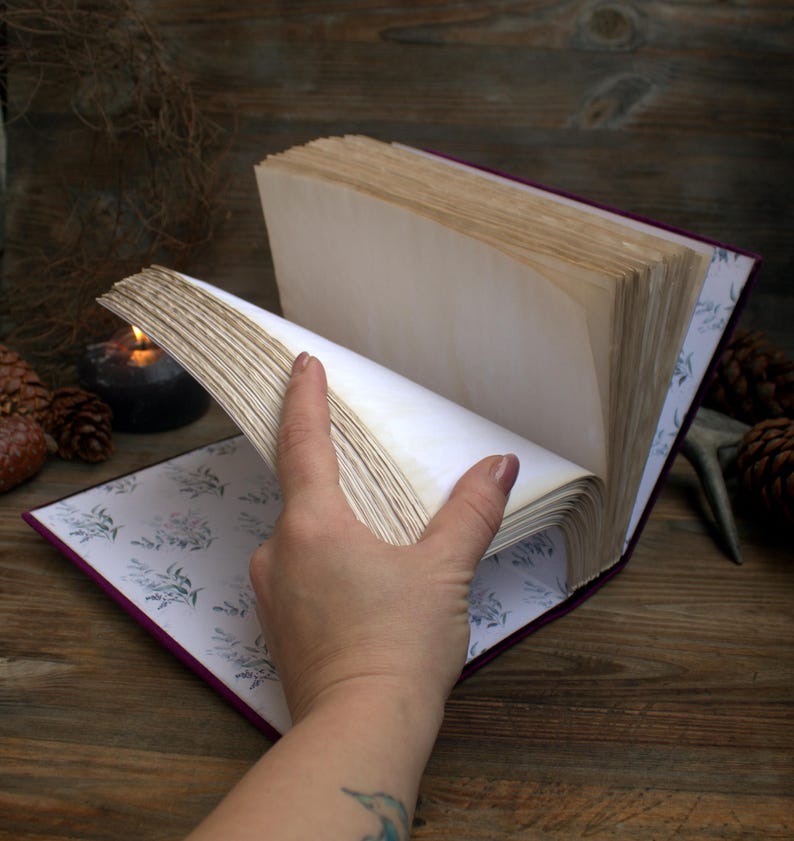 May include: An open book with aged, cream-coloured pages. The book has a deep purple cover and a floral patterned interior. A hand is turning the pages, revealing the texture of the paper. The background includes a wooden surface, pine cones, and a lit candle.