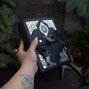 May include: Black leather box with ornate gold-colored corner accents and a central oval emblem. The box features a clasp closure and a decorative tassel. The box is held by a hand, with a tattoo visible on the arm. The box is surrounded by evergreen branches.