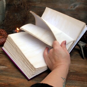 May include: An open journal with cream-coloured, aged pages. The journal has a deep purple cover and is being held open, revealing the blank pages. The image is set on a rustic wooden surface.