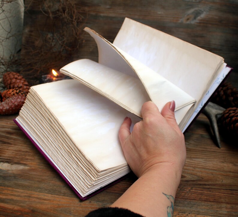 May include: An open journal with cream-coloured, aged pages. The journal has a deep purple cover and is being held open, revealing the blank pages. The image is set on a rustic wooden surface.