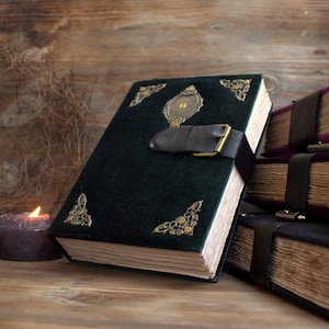 May include: A dark green velvet book with ornate corner and center accents, secured by a leather strap with a brass buckle. The book has aged, yellowed pages and is part of a collection of similar books.