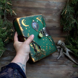 Puede incluir: Un diario verde con detalles dorados, con un diseño de luna creciente, estrellas y pájaros. El diario tiene un cierre decorativo, una llave pequeña y una correa de cuero marrón. La cubierta está adornada con adornos de metal.
