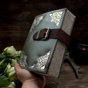 May include: A handcrafted, olive green leather-bound book with a brown leather strap and silver buckle. The corners are adorned with ornate silver filigree. The book's pages have a vintage, aged appearance.