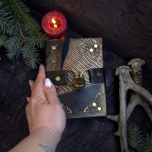 May include: A handcrafted black leather journal with gold accents and a decorative clasp. The journal features a textured gold panel and ornate corner details. A lit red candle and pine branches are in the background.