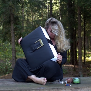 May include: A large, thick, black book with a prominent metallic buckle closure on the front cover. The book is held by a person with long blonde hair, wearing black clothing, while sitting in a forest setting with small objects nearby.