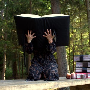 May include: A large, open black book with thick white pages held upright. Beside it sits a stack of three smaller, rectangular books bound with dark straps, and a fourth book with a red cover. The books feature visible paper edges and bindings.