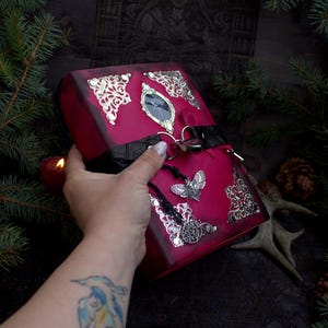 May include: A burgundy-colored, decorative box with silver filigree corners and a silver butterfly charm. The box has a black strap and a silver lock. The box is open, revealing a dark interior. The box is held by a person.