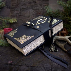 May include: A black leather-bound journal with gold filigree corner accents and a gold clasp. The journal has a black ribbon closure and a small pendant. The pages are aged, and the journal is surrounded by pine branches.