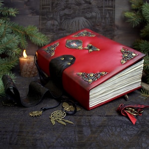 May include: A red leather-bound book with ornate corner and center accents. The book has a black leather strap closure and a decorative charm. A lit candle and pine branches are in the background.
