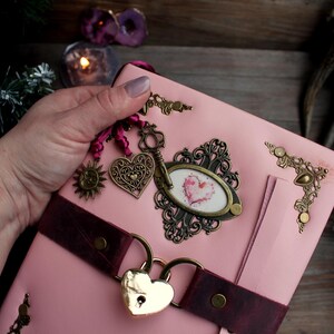 May include: Pink leather journal with a heart-shaped lock and decorative metal charms. The journal features a burgundy strap, ornate corner accents, and a framed heart design. A key charm and sun charm are also attached.