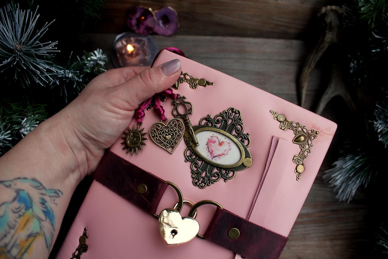 May include: Pink leather journal with a heart-shaped lock and decorative metal charms. The journal features a burgundy strap, ornate corner accents, and a framed heart design. A key charm and sun charm are also attached.