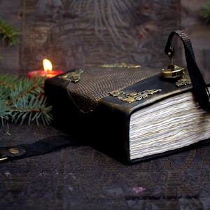Könnte beinhalten: Ein in schwarzes Leder gebundenes Tagebuch mit strukturierten Einsätzen, verzierten Messingecken und einem Metallverschluss. Das Buch hat dicke Papierseiten mit unebenen Kanten und einen schwarzen Riemen mit einem seitlichen Messingring.