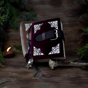 May include: A burgundy velvet journal with silver accents and a leather strap closure. The journal is sitting on a wooden surface with a candle and a twig.