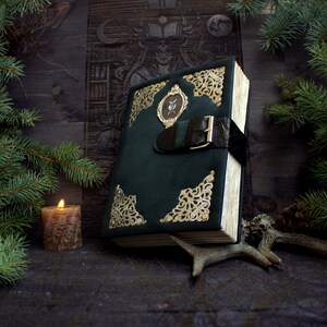 May include: A dark green leather-bound book with gold filigree corners and a central clasp. The book has a black leather strap with a gold buckle. A small oval emblem is centered on the cover. The book is propped up with a deer antler.