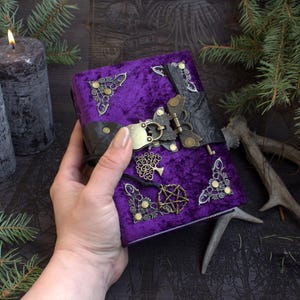 May include: A purple velvet journal featuring ornate brass corner protectors, a central black leather strap with a brass latch, and dangling charms including a tree of life and a pentagram. The cover is decorated with intricate metal accents.
