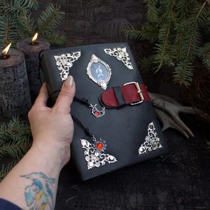 May include: A black leather-bound journal with silver corner embellishments and a red and black strap closure. The journal features a small framed image and a decorative charm with a red stone. The overall theme is gothic and fantasy.