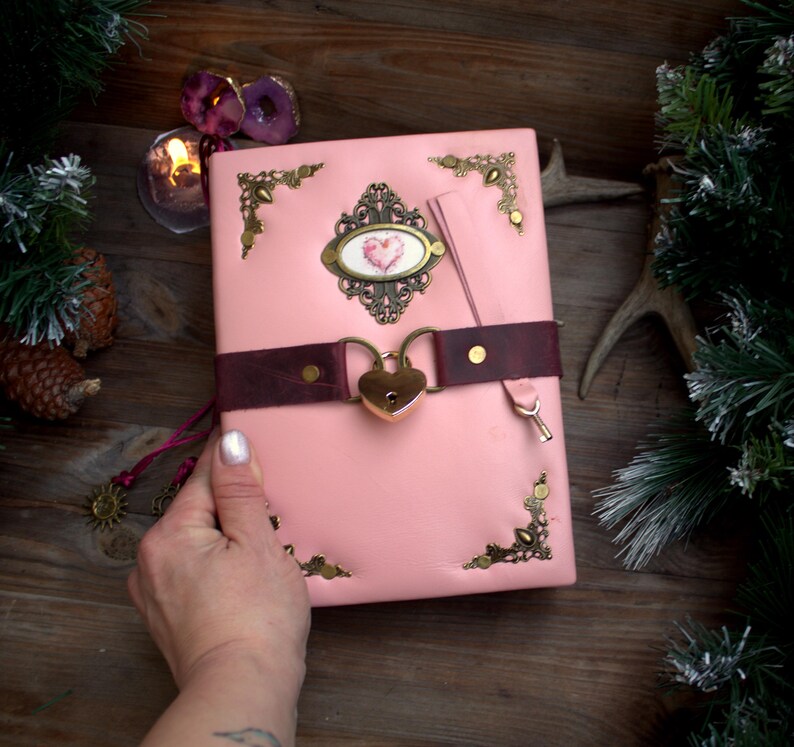May include: A pink leather journal with a heart-shaped lock and a burgundy strap. The journal features ornate corner accents and a decorative oval emblem with a heart design. A key is attached to the strap. The journal is held by a hand.