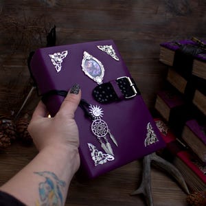 May include: A purple book with silver embellishments, including corner accents, a framed image, and a buckle closure. A silver dreamcatcher charm hangs from the book. The book is held in a hand with a tattoo and nail art. Other books are in the background.
