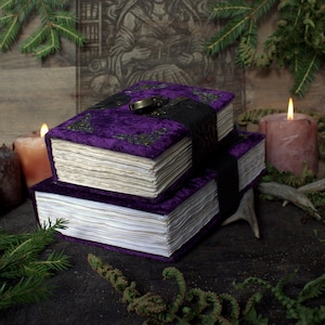 May include: Two stacked journals with deep purple velvet covers, featuring ornate metallic corner accents and a central black latch closure. The books have thick, deckled-edge paper pages and are positioned horizontally, showcasing their textured fabric.