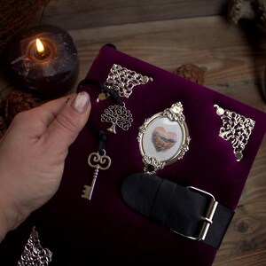 May include: A deep purple velvet journal with silver embellishments, including corner accents, a framed heart image, and a key charm. A black leather strap with a silver buckle secures the journal.