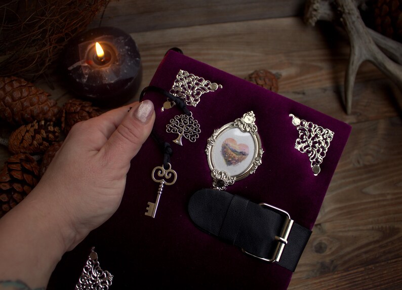 May include: A deep purple velvet journal with silver embellishments, including corner accents, a framed heart image, and a key charm. A black leather strap with a silver buckle secures the journal.