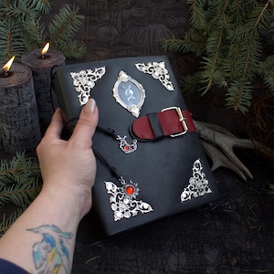 May include: A black leather-bound journal with silver corner embellishments and a red and black strap closure. The journal features a small framed image and a decorative charm with a red stone. The overall theme is gothic and fantasy.