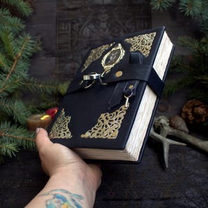 May include: A black leather-bound journal with gold filigree corner accents and a decorative clasp. The journal has a black strap closure and a gold-colored charm. The pages are aged, and the journal is held in a hand.