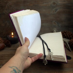 May include: A journal with a purple cover and cream-coloured pages is held open. A black cord with a key and tree charm acts as a bookmark. The open pages are displayed against a wooden backdrop, with a lit candle and pine cones.