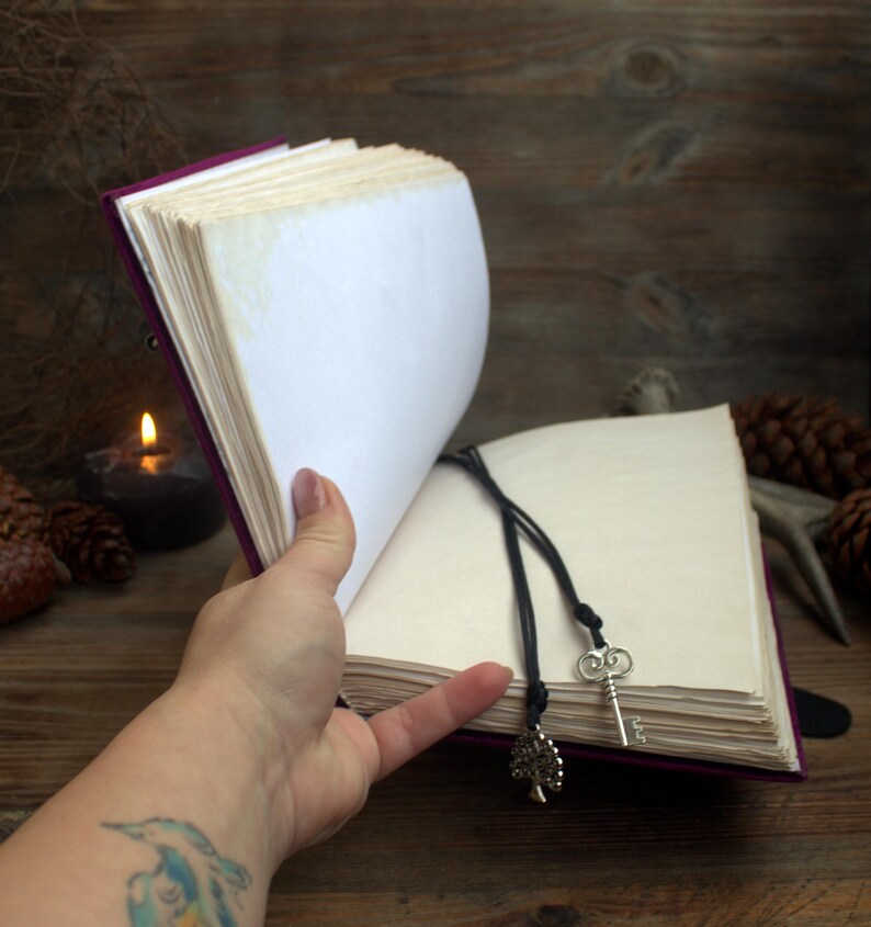 May include: A journal with a purple cover and cream-coloured pages is held open. A black cord with a key and tree charm acts as a bookmark. The open pages are displayed against a wooden backdrop, with a lit candle and pine cones.