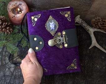 May include: A rectangular journal covered in deep purple velvet fabric, featuring ornate brass corner protectors, a central oval cabochon displaying a triple moon symbol, and a black leather strap closure secured with a decorative brass latch.