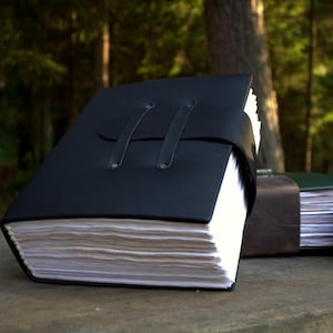 May include: A thick, black journal with a smooth cover and a wrap-around strap closure secured by two metal rivets. The journal contains a large stack of white, textured paper pages with deckled edges, visible from the side profile of the book.