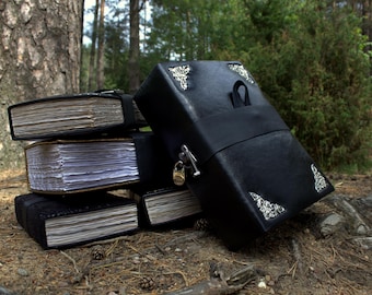 May include: A stack of black leather-bound journals and a larger book-shaped box with silver corner accents. The journals have aged, textured pages. The box has a lock and strap closure, set outdoors.