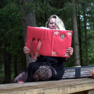 May include: A bright coral red book with a leather-like cover, featuring a brown strap handle, metallic gold corner accents, and a central eye-shaped embellishment. The book is held open, revealing white pages, while a second black book sits nearby.